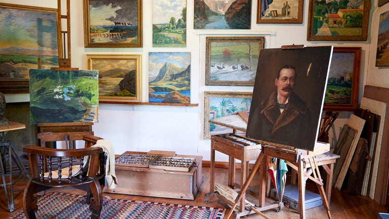 Interior of Winston S. Churchill's studio at Chartwell, Kent. Paintings by Churchill displayed on walls. Box of paints on the floor. Chair draped with a beige jacket sits in front of and easel with painting of the golden orfe pond. Another portrait stands on an easel.